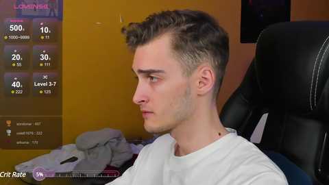 Media: Video of a young, fair-skinned, brown-haired man in a white shirt, seated in a black gaming chair, indoors with a messy bed in the background.