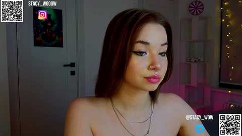 Media: A video of a young, fair-skinned woman with straight, shoulder-length brown hair, wearing a nose ring and necklace. She's topless, indoors, with a colorful wall art behind her.