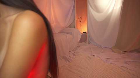 Media: Video of a dimly lit bedroom with white, sheer curtains draped over the bed. A soft, warm light emanates from the background, creating a cozy, intimate atmosphere. The room has a minimalist, serene feel.