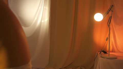 Media: Video of a dimly lit, cozy bedroom with warm orange hues. White curtains are drawn, casting a soft glow on the room. A small, white lamp on a bedside table emits a gentle light.