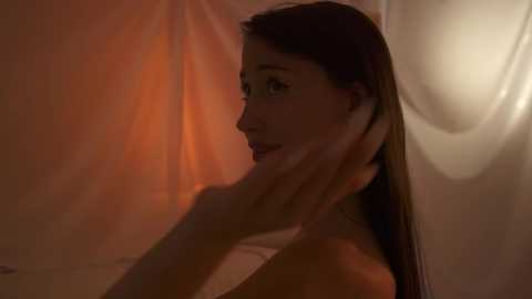 Media: A video of a young woman with long, straight brown hair, looking contemplative. She's partially nude, with her hand gently touching her cheek. The background is softly lit, featuring warm, orange hues and a blurred curtain.