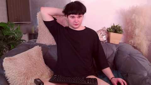 Media: Video of a young man with fair skin and black hair, wearing a black t-shirt, sitting on a gray couch with beige pillows, holding a black keyboard.