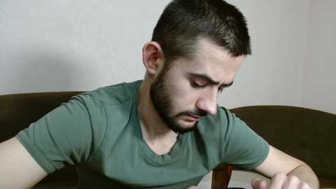 Media: Video of a young man with short dark hair, beard, and olive-green T-shirt, leaning forward on a brown couch, looking down, in a plain, beige-walled room.