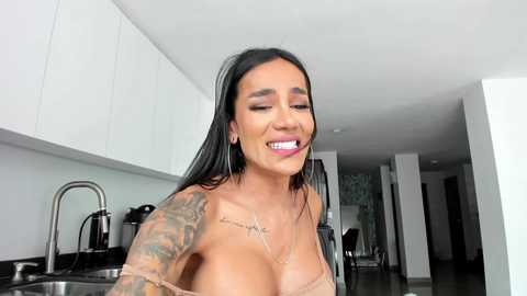 Media: Video of a smiling, tattooed woman with medium brown skin, black hair, and a revealing beige top, standing in a modern kitchen with white cabinets and stainless steel appliances.