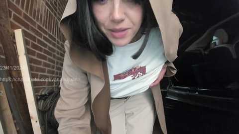 Media: Video of a young woman with shoulder-length black hair, wearing a beige trench coat and a light blue shirt, bending over in a dimly lit garage.