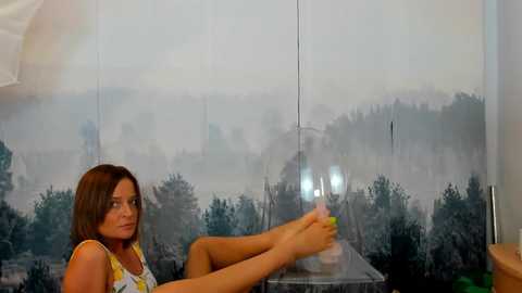 Media: Video of a woman with shoulder-length brown hair, wearing a yellow top, sitting on a window sill with legs extended, looking out at a misty landscape.