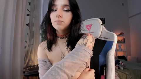 Media: Video of a young woman with pale skin, dark hair, and a tattoo sleeve, wearing a sheer top, sitting in a gaming chair in a dimly lit room.