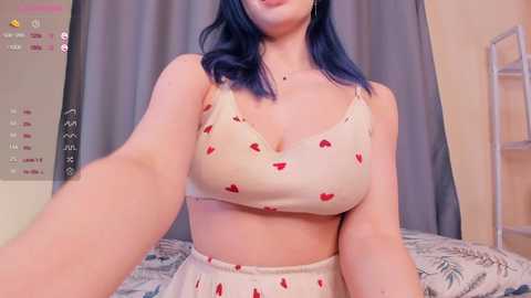 Media: Video of a woman with blue hair in a beige crop top and matching pants, taking a selfie in a bedroom with a calendar and gray curtains.