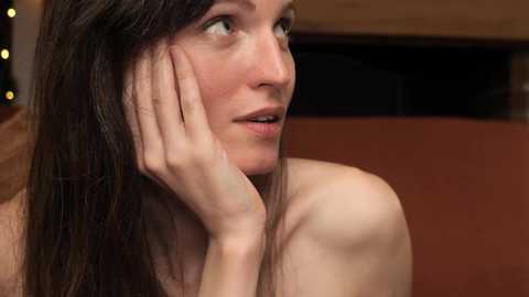 Media: Video of a Caucasian woman with long, dark hair, fair skin, and freckles, looking introspective, holding her cheek with her hand. She's bare-shouldered, possibly in a living room with warm lighting and a wooden backdrop.