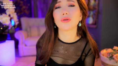 Media: Video of a young woman with long brown hair, wearing a black mesh top and dangling earrings, set against a dimly lit room with floral arrangements and a white sofa in the background.