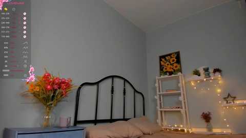 Media: Video of a cozy, minimalist bedroom with light gray walls, black wrought-iron headboard, beige bedding, floral arrangement, star-shaped decorations, and a wooden ladder shelf with fairy lights.