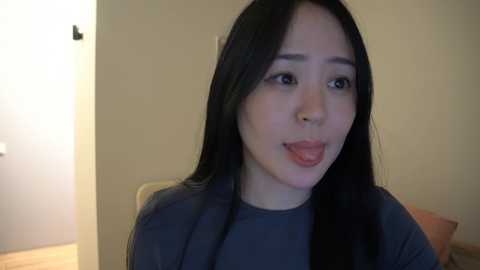 Media: A video of an Asian woman with long black hair, wearing a dark top, playfully sticking her tongue out, in a dimly lit indoor setting with beige walls and a wooden floor.