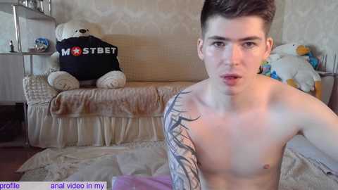 Media: Video of a shirtless young man with short brown hair, tattoos, and pale skin, sitting on a beige couch with plush toys and a \"MST3K\" shirt, in a cluttered living room.