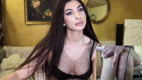 Media: Video of a young woman with long, straight black hair, wearing a black mesh top, and sitting on a beige couch in a cozy, warmly-lit living room.