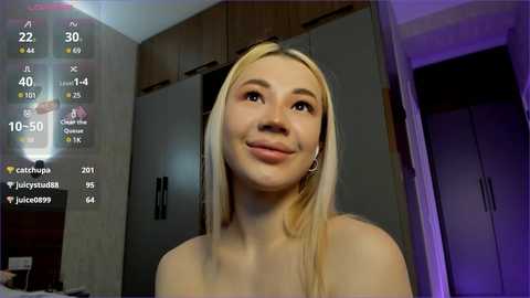 Media: Video of a smiling young woman with long blonde hair and fair skin, standing in a modern, dimly-lit bedroom with gray cabinets and purple lighting. Social media icons and a digital clock display are overlaid.