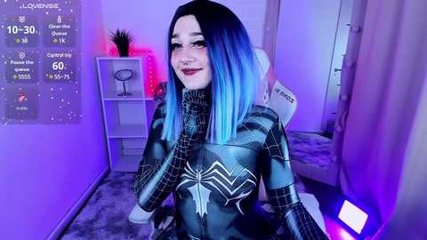 Media: Video of a young woman with light skin, blue-black hair, and a slender physique, dressed in a Spider-Man costume, sitting on a grey carpeted floor in a dimly lit room with purple lighting.