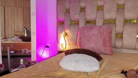 Media: Video of a cozy bedroom with a plush, light pink headboard, a gold-framed mirror, and a soft white pillow on a beige bedspread. Ambient lighting casts a warm purple glow.