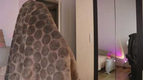 Media: A video of a person in a fuzzy, gray, circular-patterned blanket standing in a modern bedroom with a mirrored closet door, a bed, and purple lighting.