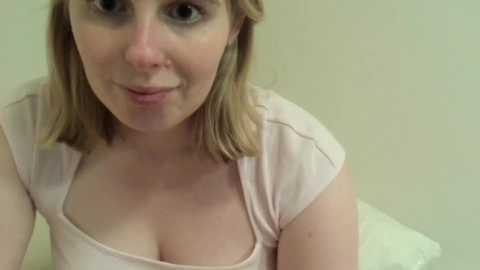 Media: Video of a fair-skinned, blonde woman with shoulder-length hair, wearing a pink top that reveals cleavage, sitting against a light-colored wall. She has a slight smile.