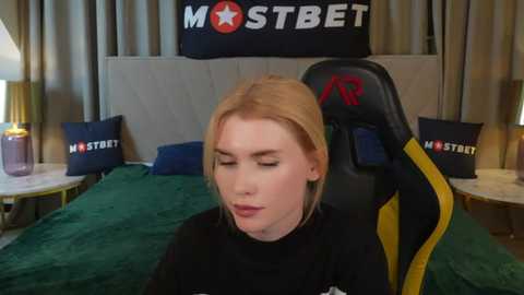 Media: Video of a blonde woman in a black shirt, seated in a yellow and black gaming chair in a bedroom with a green bedspread, beige curtains, and \"MostBet\" decor.