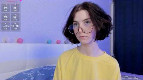 Video of a young woman with short brown hair, wearing glasses and a yellow shirt, standing in a brightly lit room with a white bed and colorful decorative balls in the background.