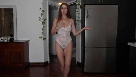 Media: Video of a slender, fair-skinned woman with long red hair, wearing a lacy, sheer white bodysuit, standing in a modern kitchen with a stainless steel refrigerator and dark countertops.