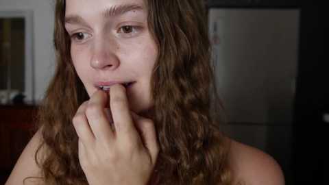 Media: A video of a young woman with long, wavy brown hair, pale skin, and a thin build, biting her finger, standing in a dimly lit kitchen with a silver refrigerator in the background.