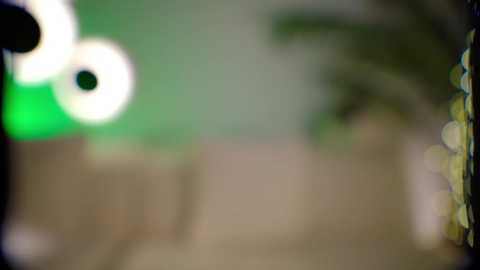 Media: Video with a blurred background, featuring a green and white light source on the left, a beige wall in the middle, and a blurry green plant on the right. The overall image is soft and out-of-focus.