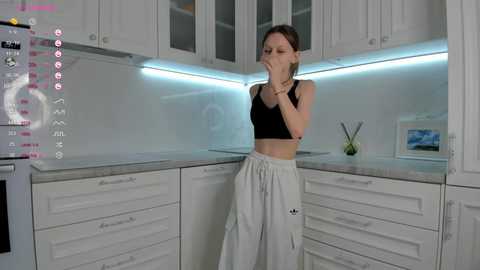 Media: Video of a slender woman in a black sports bra and white Adidas pants, covering her mouth in a modern kitchen with white cabinets, blue LED strip lighting, and minimal decor.