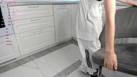 Media: Video of a woman in white Adidas tracksuit, standing in a modern kitchen with white cabinets, grey chairs, and tiled floor, looking at a calendar on the wall.