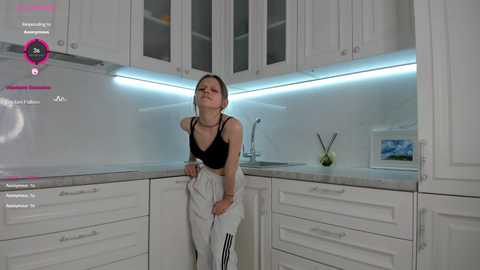 Media: Video of a young Caucasian woman in a sleek, white kitchen. She's wearing a black sports bra and gray sweatpants, with a slight smile. Background includes modern white cabinets, sleek countertops, and a blue LED strip light.