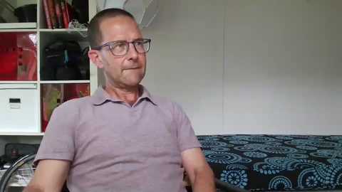 Media: Video of a middle-aged man with glasses, balding, wearing a light purple polo shirt, seated in a cluttered bedroom with white shelves and a bed covered with a blue-patterned blanket.