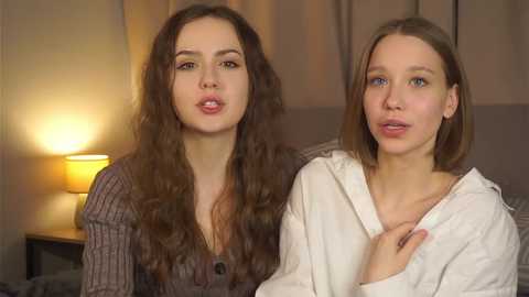 Media: Video of two young women, one with long curly hair in a ribbed sweater, the other with straight hair in a white shirt, standing close in a warmly-lit room with a lamp.