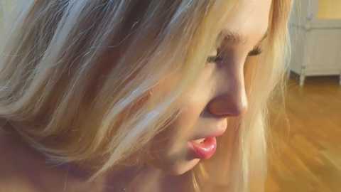 Media: Close-up video of a young Caucasian woman with long, wavy blonde hair, fair skin, and red lipstick. She's looking downward, her face partially in shadow. Background shows a wooden floor and blurred, light-colored walls.