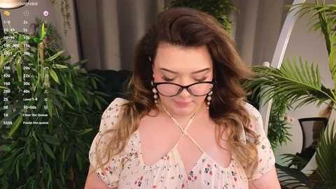 Media: Video of a fair-skinned woman with wavy brown hair, wearing glasses, a floral dress, and a pearl necklace, standing in a lush indoor setting with greenery.