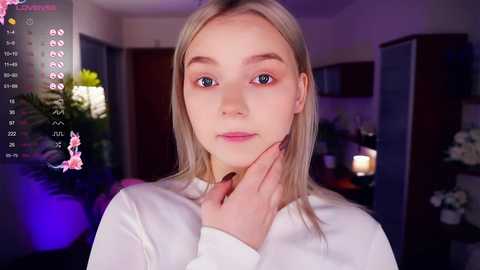 Media: Video of a young, light-skinned woman with straight blonde hair, wearing a white top, indoors with a blurred background featuring green plants and candles.