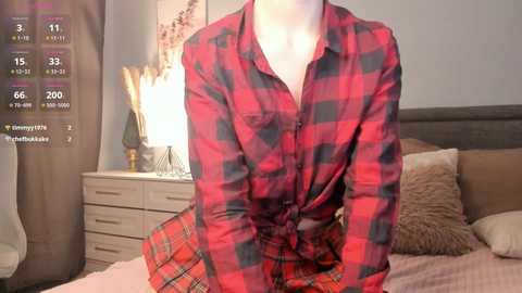 Media: Video of a woman in a red and black plaid shirt, sitting on a bed with a beige headboard, in a cozy bedroom setting.