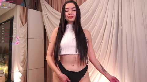 Media: A video of a slender, light-skinned woman with long black hair, wearing a white crop top and black panties, standing confidently in a dimly lit room with beige curtains and a soft, diffused lighting.