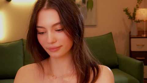 Media: Video of a young woman with straight, shoulder-length brown hair, fair skin, and a delicate face. She wears a light, off-the-shoulder top. Background features a green sofa and a warm, cozy room with a potted plant and framed artwork.