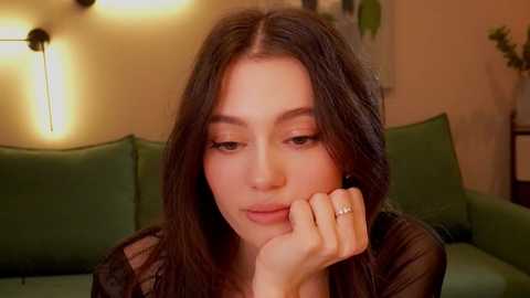 Media: A video of a young woman with long, dark hair and fair skin, wearing a black sheer top, resting her chin on her hand, in a cozy, warmly lit living room with green couches and potted plants.