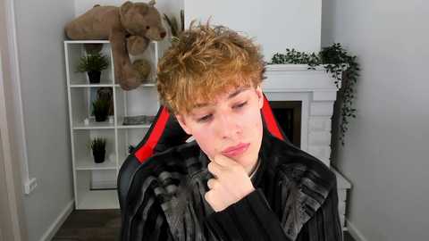 Media: A video of a young boy with curly red hair, wearing a black jacket, sitting on a red gaming chair, contemplating with his chin on his hand. Background includes a white fireplace, a plant, and a stuffed bear.