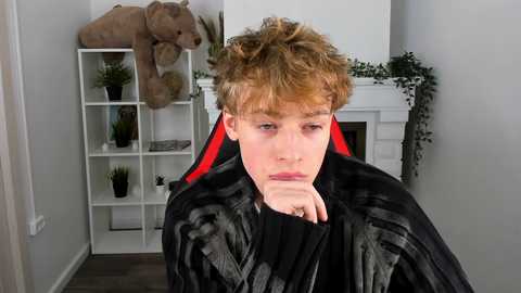 Media: Video of a young, pale-skinned, curly-haired boy in a black cape with red lining, looking pensive, in a minimalist, modern living room with white shelves and a large elephant toy.