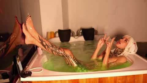 Media: Video of a nude woman with long white hair and tattoos, reclining in a green-tinted hot tub, surrounded by black cushions.