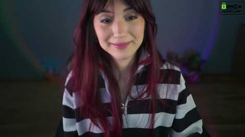 Media: Video of a young woman with long, straight, dark red hair, wearing a black and white striped hoodie, smiling softly in a dimly lit room with blurred background.