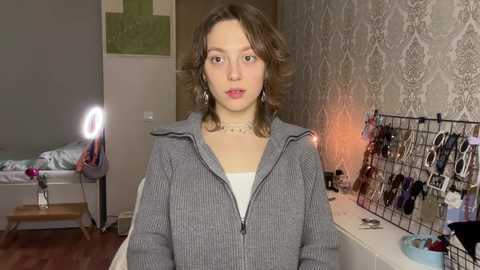 Media: Video of a young woman with shoulder-length wavy brown hair, wearing a gray zip-up hoodie, standing in a bedroom with patterned wallpaper, a bed, and a makeup mirror.