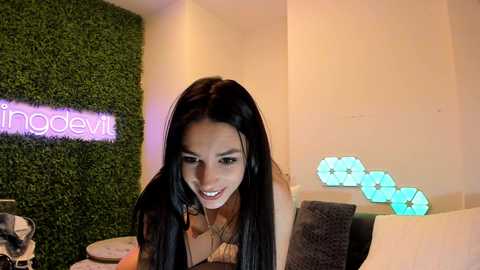 Media: Video of a young Asian woman with long black hair, wearing a brown top, smiling at the camera in a modern, green-foliage wallpapered room with blue LED lights.