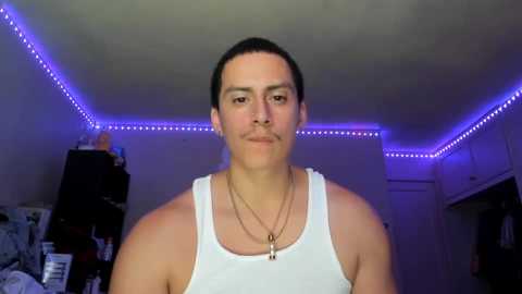 Media: Video of a light-skinned, muscular man with short dark hair, wearing a white tank top and multiple gold necklaces, standing in a dimly-lit, purple-lit bedroom with a messy closet and bed.