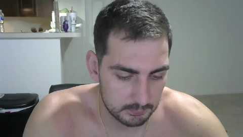 Media: Video of a shirtless man with short dark hair and a beard, sitting indoors. Background shows a kitchen counter with bottles and a sink.