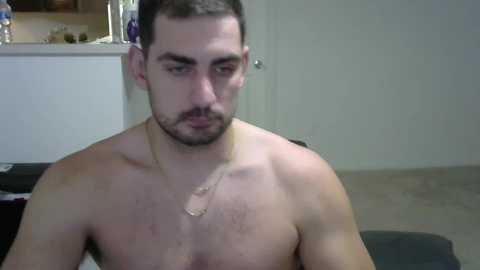 Media: Video of a shirtless, bearded, light-skinned man with short dark hair and a bandage on his chest, sitting indoors. Background features a white door, beige floor, and a kitchen counter with bottles.