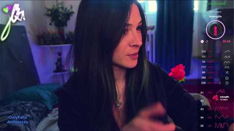 Media: A video of a woman with long, dark hair, wearing a black jacket, sitting indoors. Background shows a messy room with a bed, a TV, and a red rose.